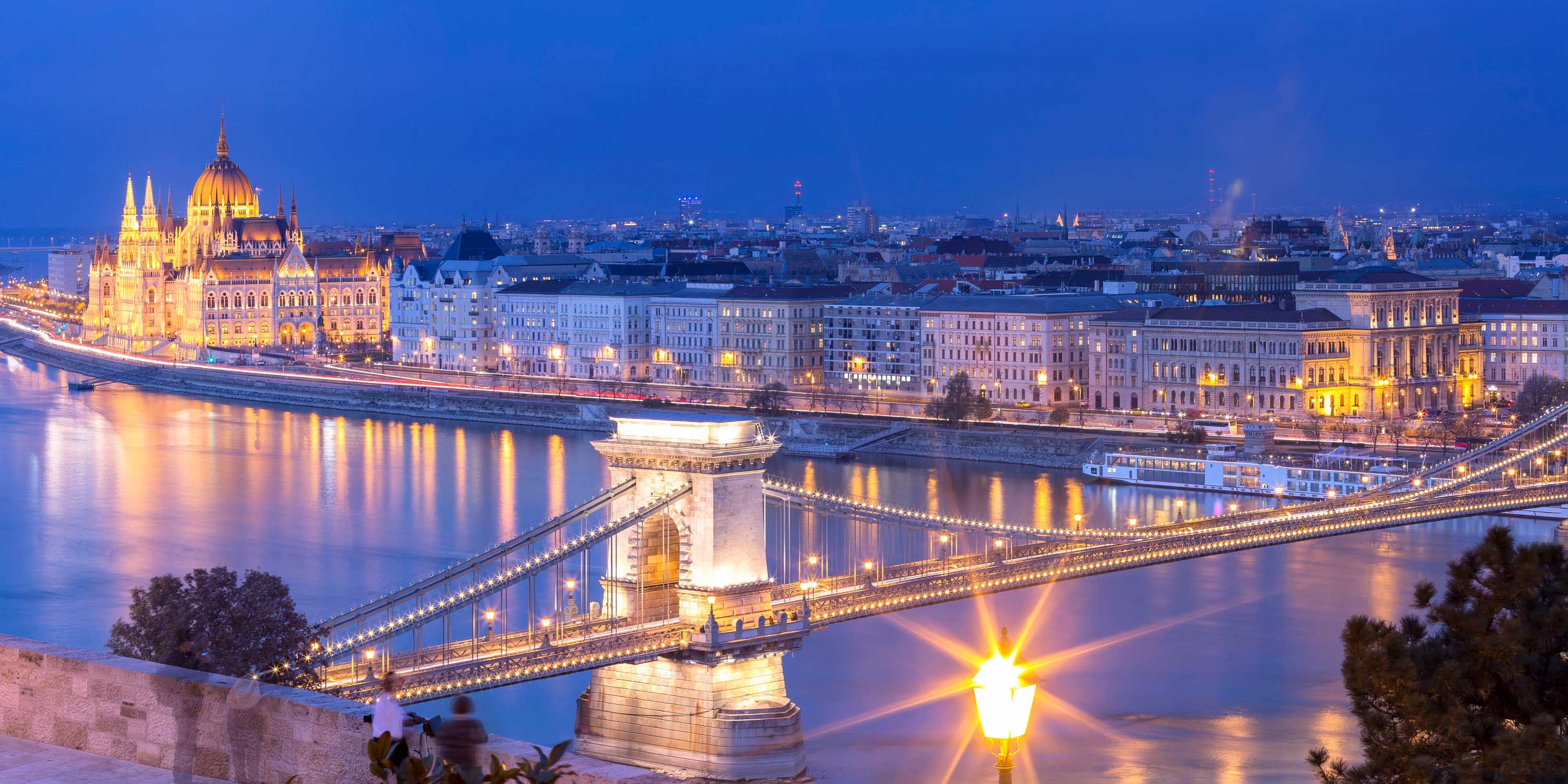A panorama of Budapest and the Danube River lit up by street and building lights in the evening