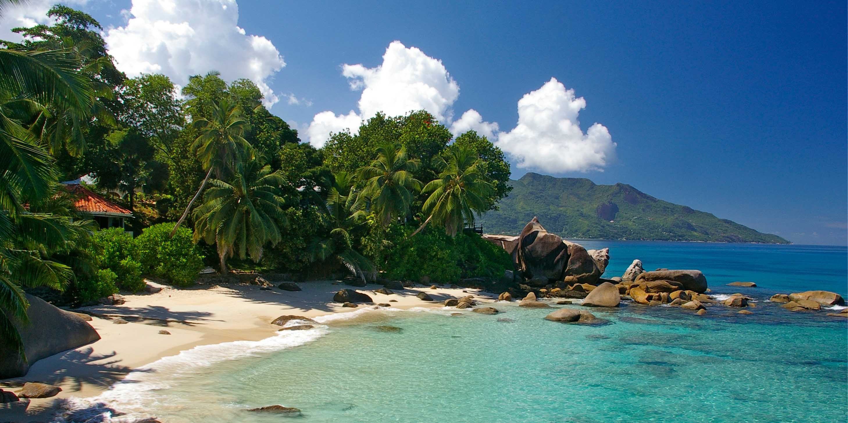 lush green foliage lining a white sandy beach with rocks scattered in the water, turquoise clear blue water and  a mountainous terrain in the background