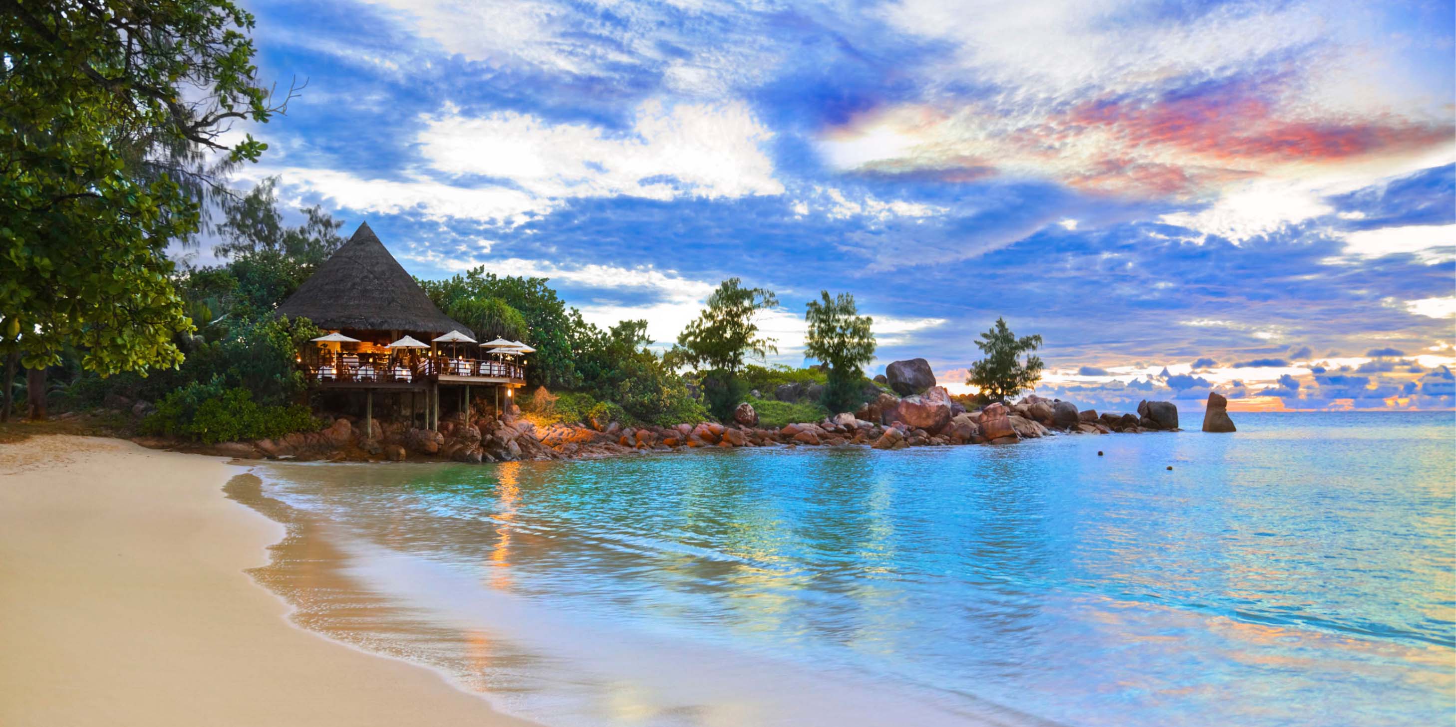 island thatched roof hut on the edge of a sandy beachfront with crystal blue water at dust with a sunset in the sky