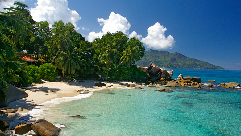 lush green foliage lining a white sandy beach with rocks scattered in the water, turquoise clear blue water and  a mountainous terrain in the background