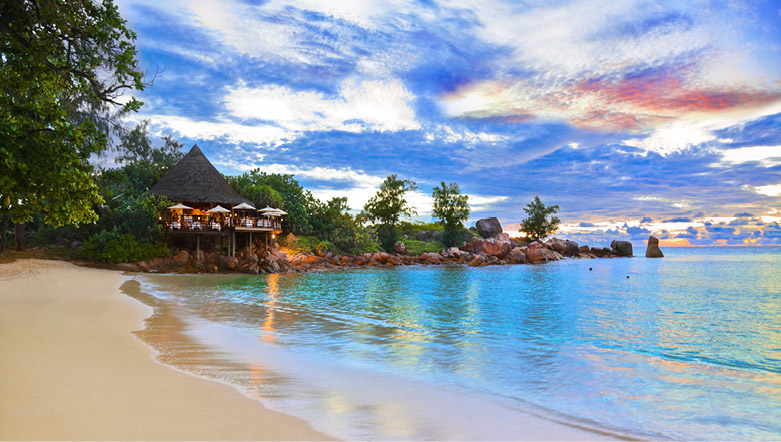 island thatched roof hut on the edge of a sandy beachfront with crystal blue water at dust with a sunset in the sky
