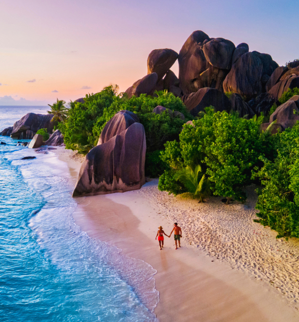 Arial view of the La Digue island in the Seychelles showing a couple walking along the beach shore lined with dark cliffs and greenery. 