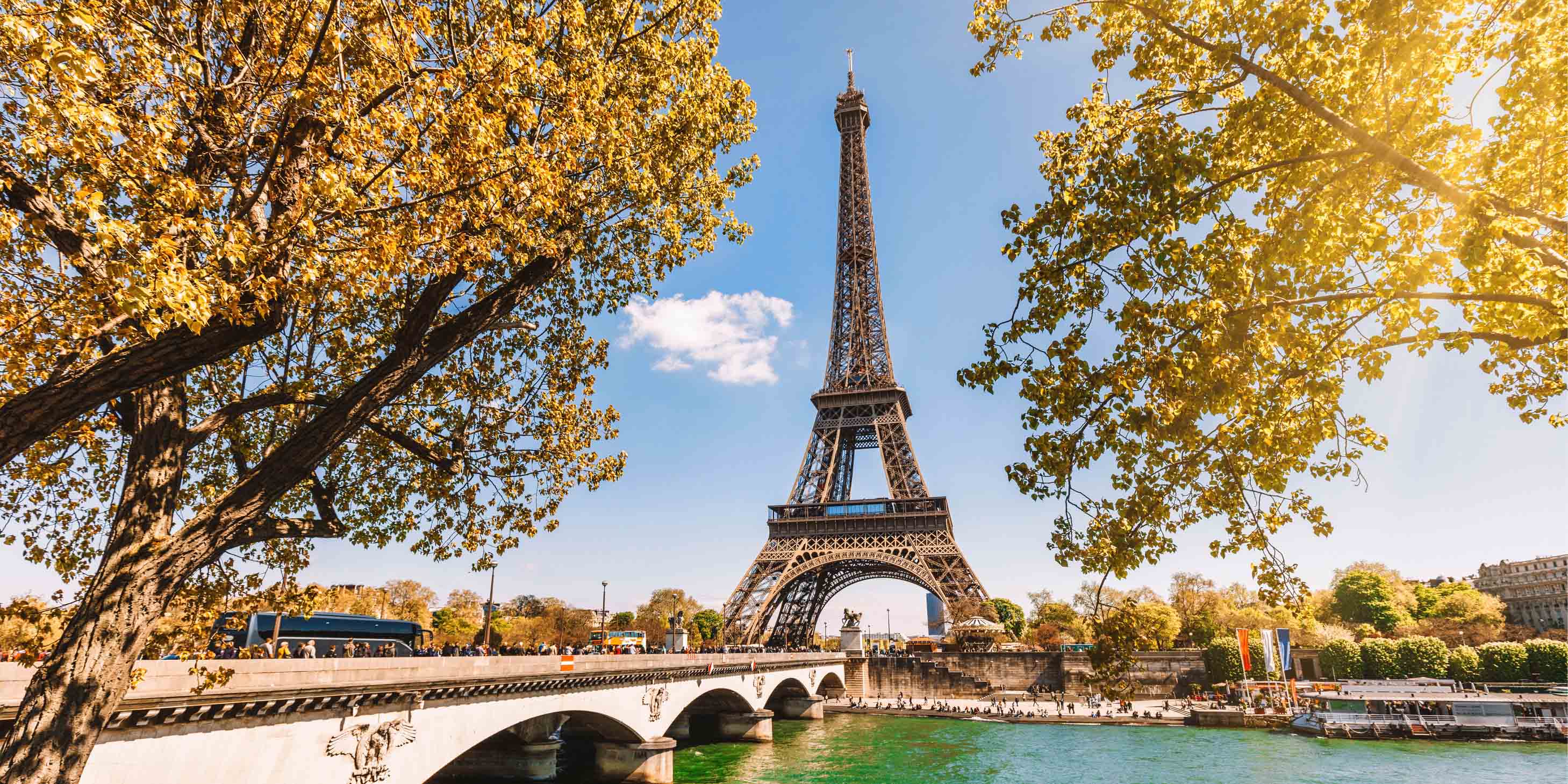 Eiffel Tower on a sunny day, seen from the Seine River, with two trees and Pont d’léna in the foreground