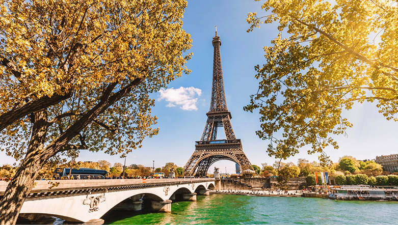 Eiffel Tower on a sunny day, seen from the Seine River, with two trees and Pont d’léna in the foreground