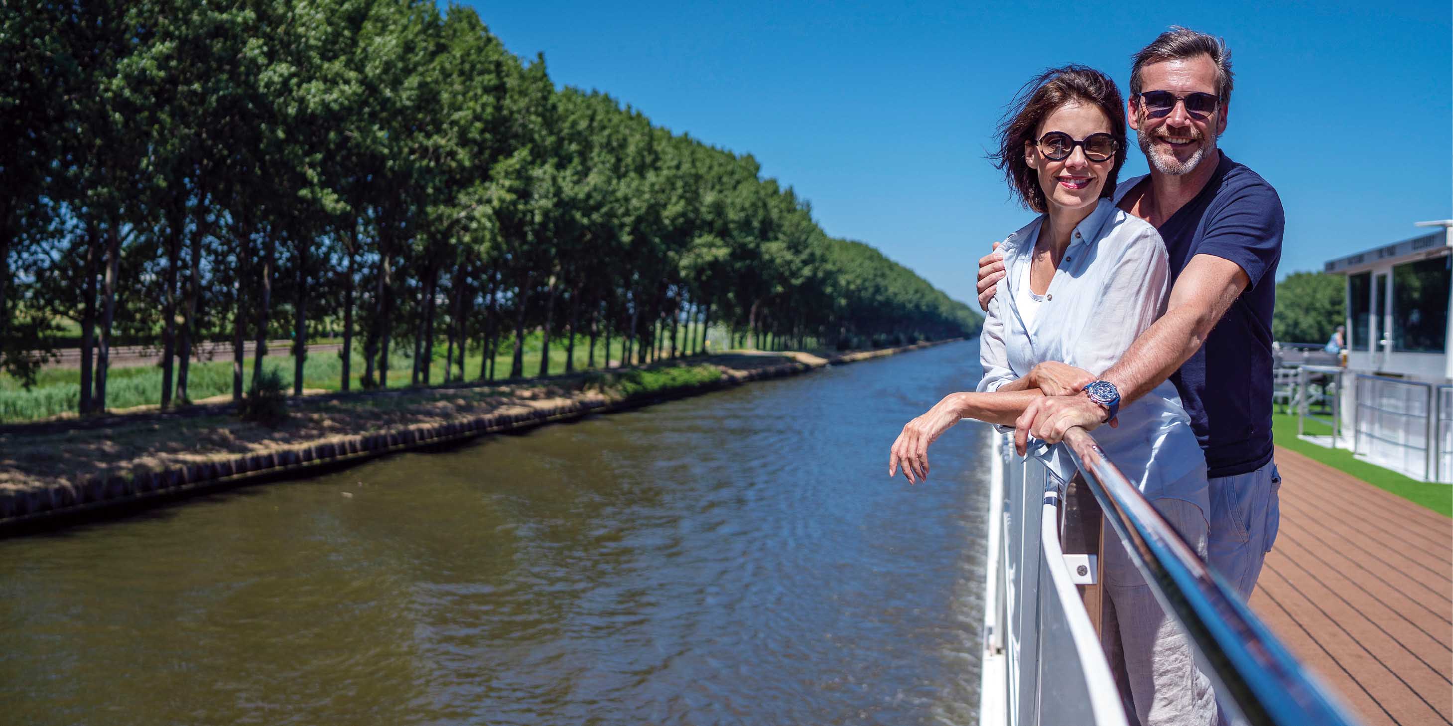 Smiling couple on the top deck of a cruise ship, sailing a river past green trees under a bright blue sky