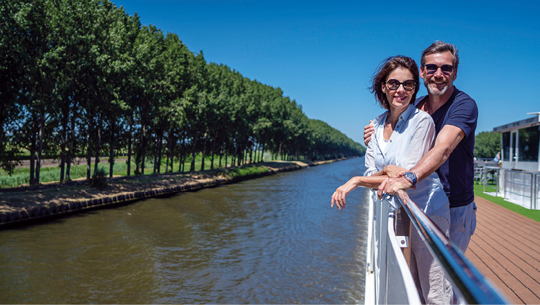 Smiling couple on the top deck of a cruise ship, sailing a river past green trees under a bright blue sky