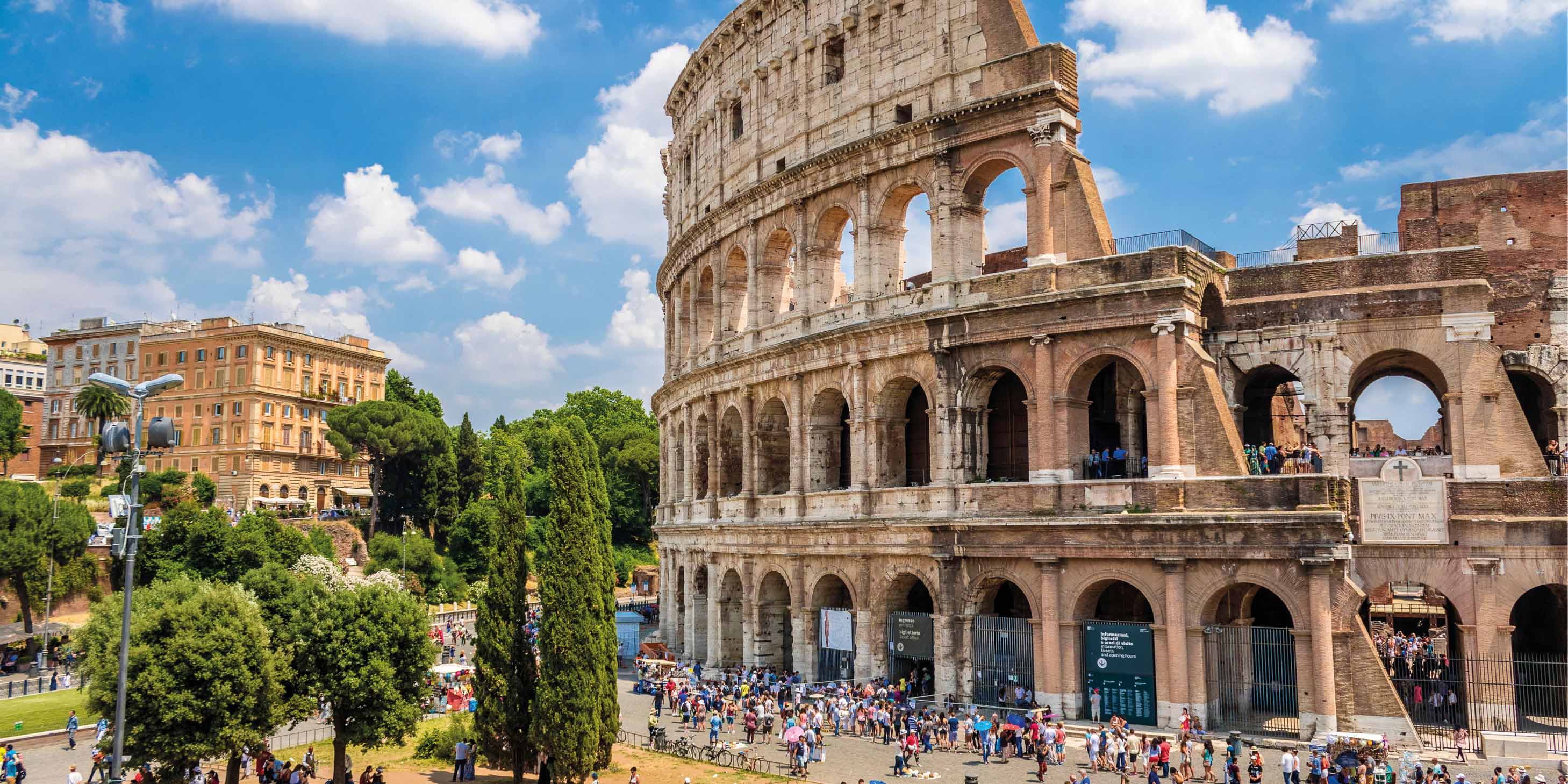 A side view of the Colosseum in Rome, with people lined up outside under cloudy blue skies on a sunny day
