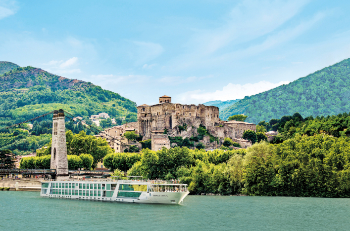 A river cruise ship passing by on a calm body of water, in front of a huge castle englufed by greenery in France. 