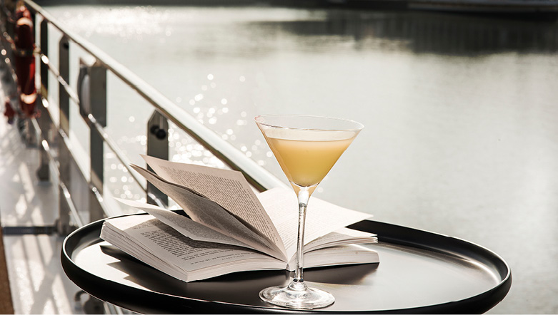 An open book next to a cocktail on a table on a cruise deck, overlooking a river, with a cruise ship in the background