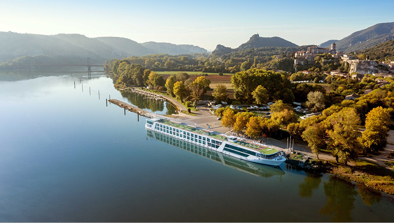 An Emerald Cruises Star-Ship is docked in Viviers, France, Green hills and a castle are seen in the background.
