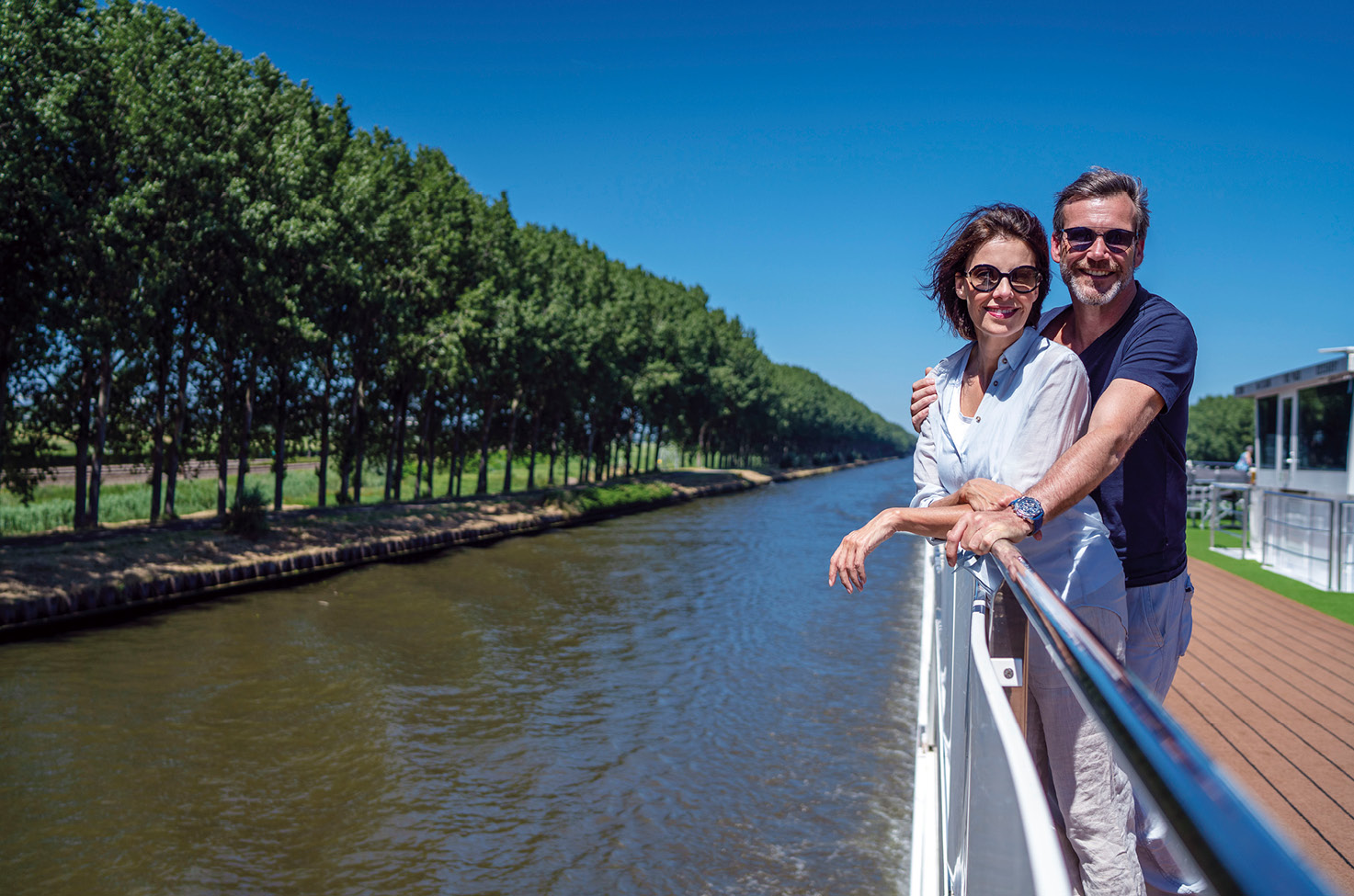 Two guests enjoying the river breeze from the top deck of an Emerald Cruises river ship as they sail through the picturesque countryside and tree-lined banks of the river