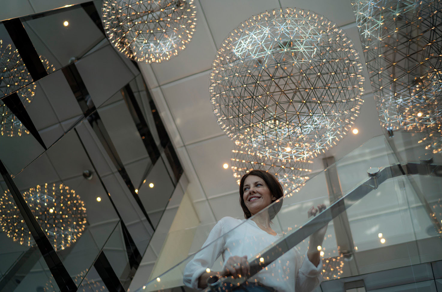 A woman on the stairs looking over the balcony and smiling, with stunning ornate lighting hanging from the ceiling