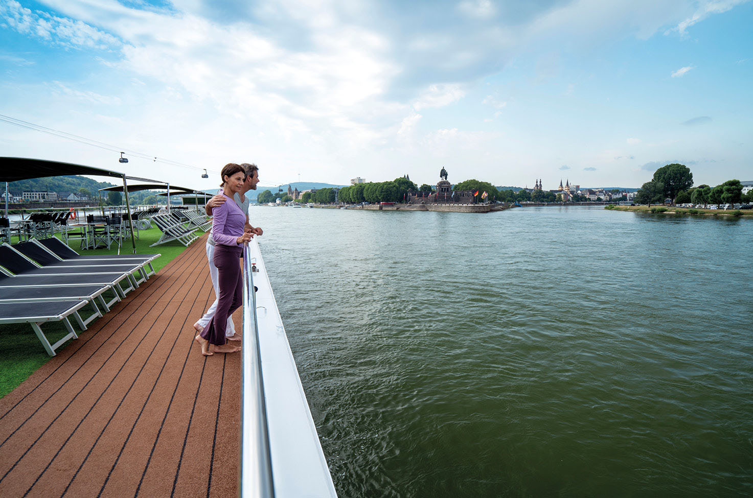 A man and woman close together on the top deck of a cruise ship, looking out to the water and the nearby town