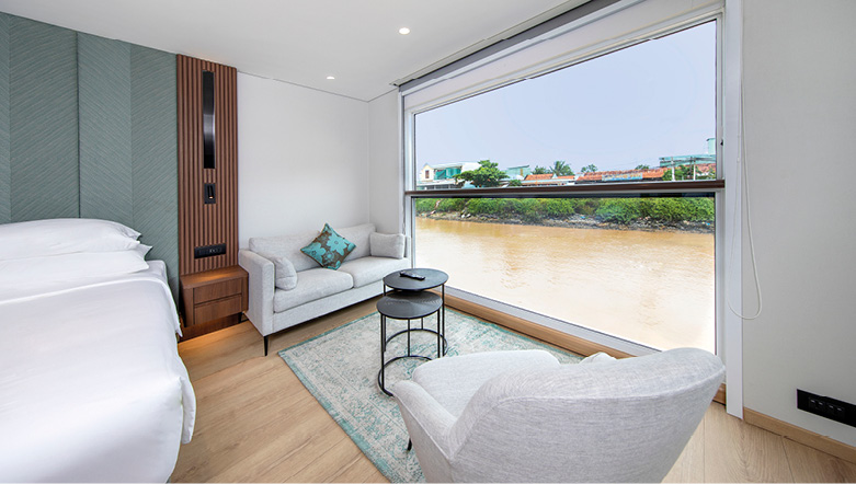A two-seater sofa and chair next to a large balcony with views of the Mekong River, with a crisp, white bed to the side of the room