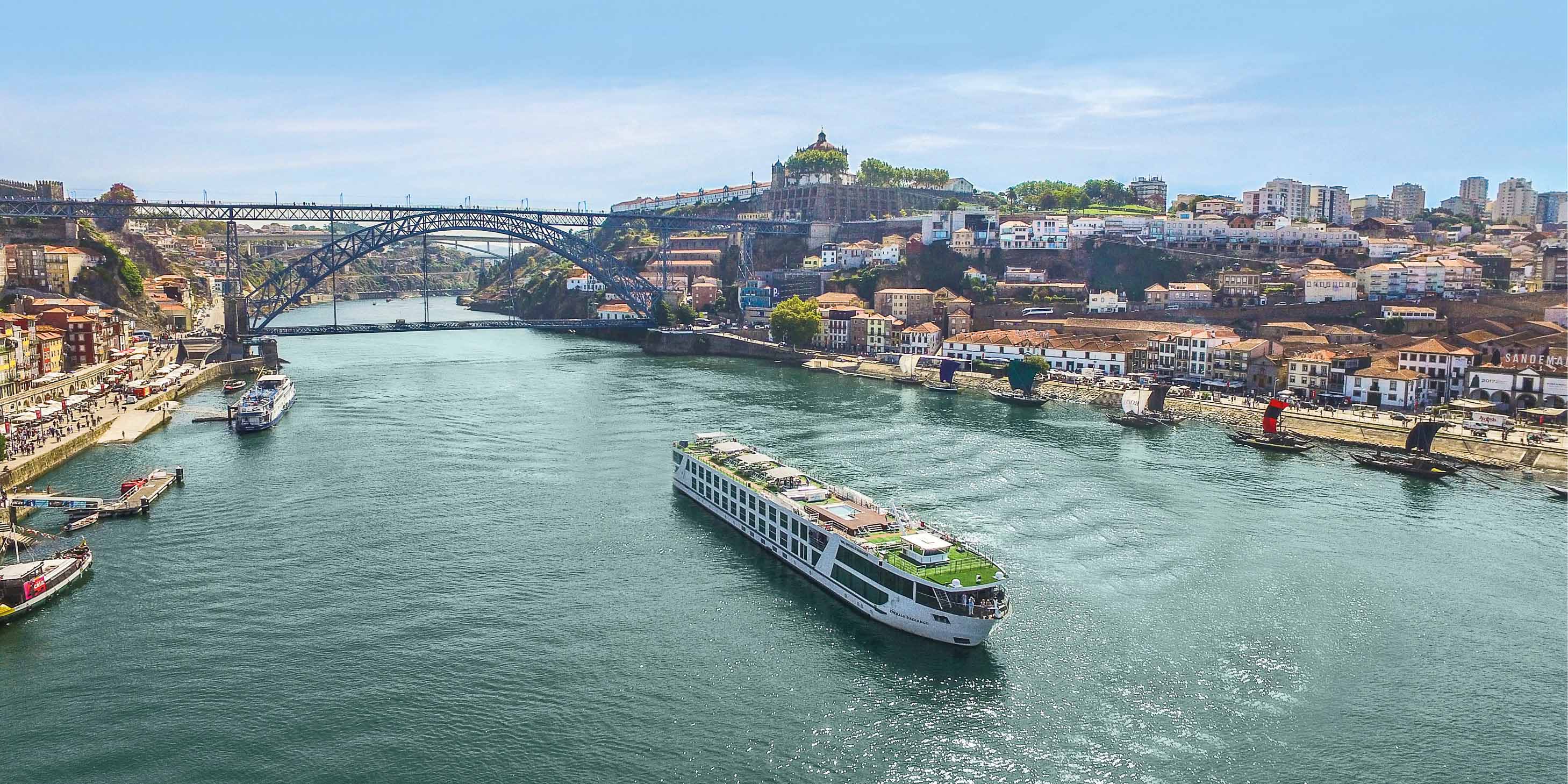 River cruise ship sailing in the middle of a river with the city of Porto on either side and an arched bridge in the background.