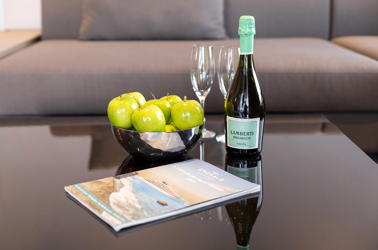 Glossy table with a bowl of green apples, a bottle of prosecco and glasses with an Emerald Cruise yacht cruises brochure