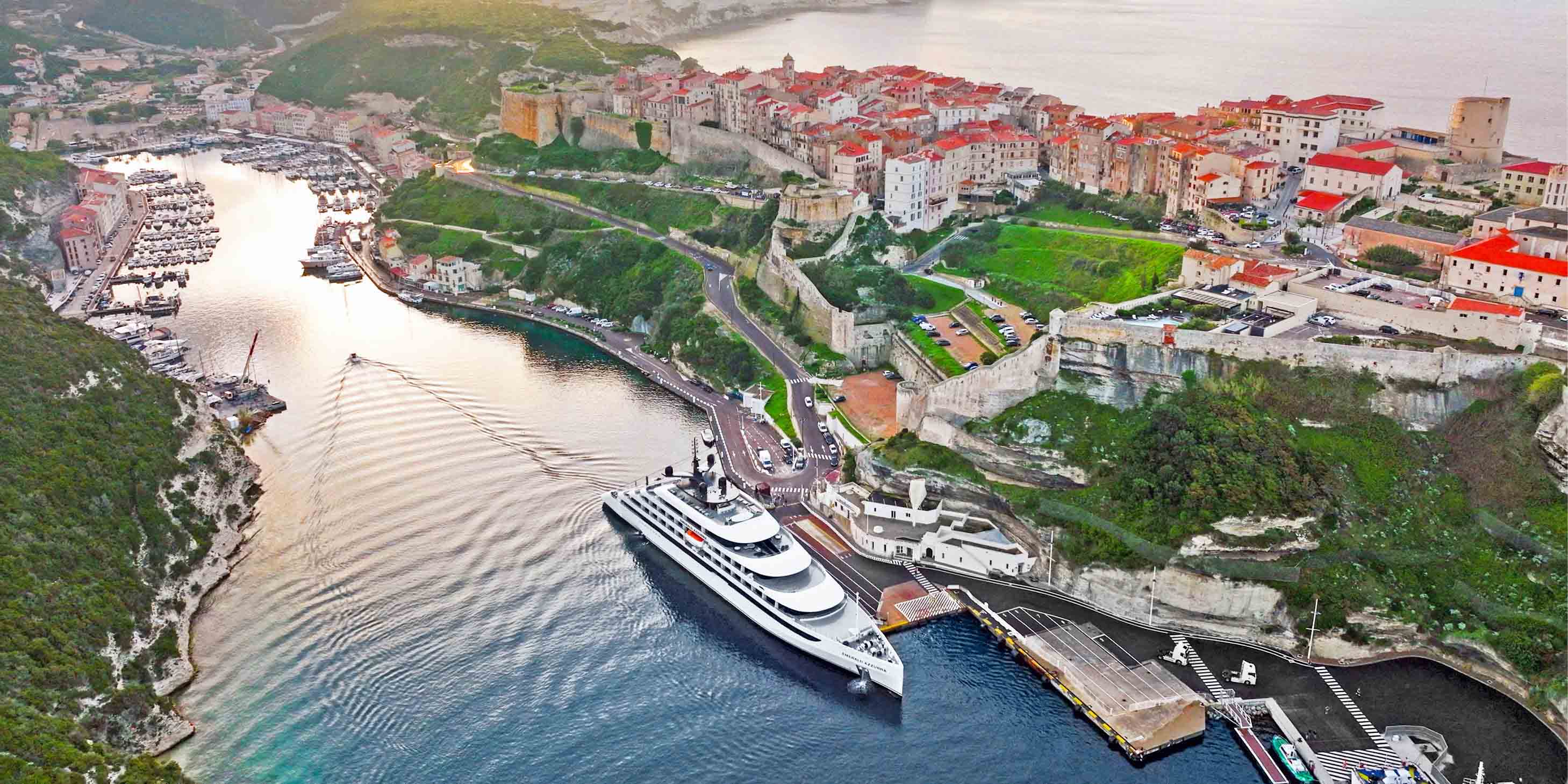 An Emerald Cruises yacht is docked in the heart of Bonifacio, Corsica, with terracotta roofs seen in the background.