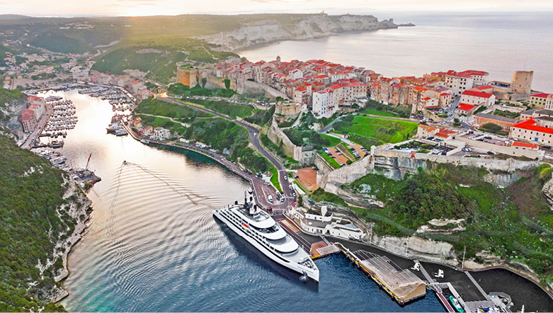 An Emerald Cruises yacht is docked in the heart of Bonifacio, Corsica, with terracotta roofs seen in the background.