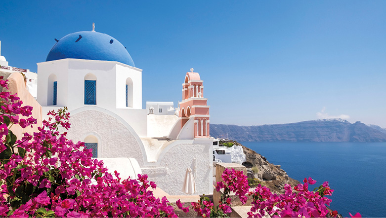 Blue-domed church of Santorini on a bright, sunny day, with the water and sky in background, and flowers in foreground    
