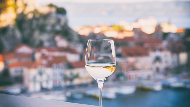 A glass of white wine in the foreground of a Mediterranean port town, which is in the shade