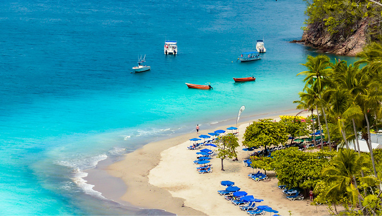 Small fishing boats in the crystal-clear blue waters and sunbeds on a palm tree lined white sandy beach in Tortuga in the Caribbean